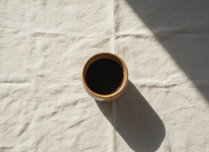 Minimal warm overhead flat-lay of a single ceramic cup of black coffee on a cream linen surface with soft morning light and gentle shadows, refined premium specialty coffee aesthetic, generous negative space, no text
