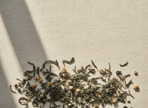 Cinematic full-bleed editorial photograph of loose-leaf artisanal tea, muted deep green tea leaves and dried botanicals arranged on a warm cream linen surface with soft directional daylight and long shadows, minimalist high-fashion still life, earthy sophisticated tones of charcoal, sage green and cream, generous negative space, luxury tea brand aesthetic like Aesop, no text, no hands, refined and quiet