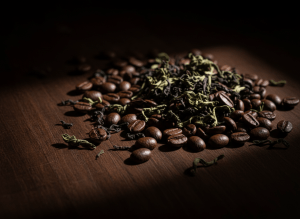 Moody cinematic close-up of dark coffee beans and loose tea leaves scattered on a dark espresso-brown surface, dramatic low-key side lighting, deep shadows, editorial food photography, extreme high contrast, no text, no people, premium specialty coffee brand aesthetic