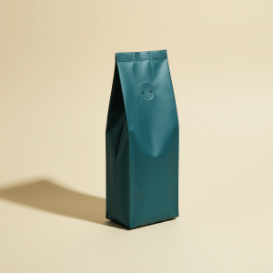 Premium matte deep teal coffee bag package standing upright on a solid warm cream background, minimalist mid-century product photography, soft directional shadow, no text or logos, editorial studio lighting