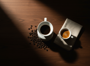 Premium editorial coffee flat lay. Wide landscape, full-bleed. Top-down overhead view on a rich dark espresso walnut wood surface. A white ceramic pour-over dripper mid-brew, freshly roasted whole coffee beans scattered naturally around it, a sleek white espresso cup with rich golden crema, soft cream linen napkin folded neatly beside it. Dramatic moody side lighting from the left, deep natural shadows. Deep espresso brown, black, warm amber, and cream tones. No text, no people, no logos, no watermarks. Shot like a Kinfolk or Monocle magazine spread. Ultra high resolution, cinematic depth of field.