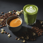 Ultra-premium editorial hero photograph: an elegant flat lay on dark slate, featuring a perfectly poured espresso in a ceramic cup with rich crema, alongside a delicate glass of vibrant green matcha latte with foam art, surrounded by scattered coffee beans and dried jasmine tea flowers. Moody, cinematic lighting with deep shadows, golden highlights. Shot for a luxury lifestyle magazine. Photorealistic, 8K quality.